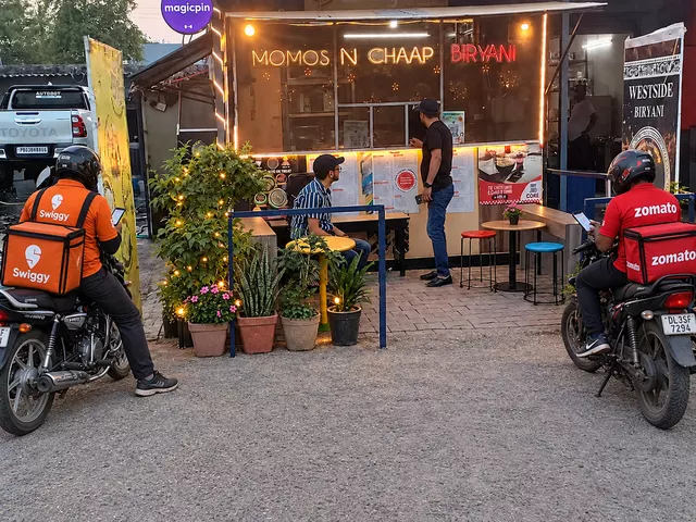 Momos N Chaap counter with privacy-safe customers and two delivery riders waiting at the frontage